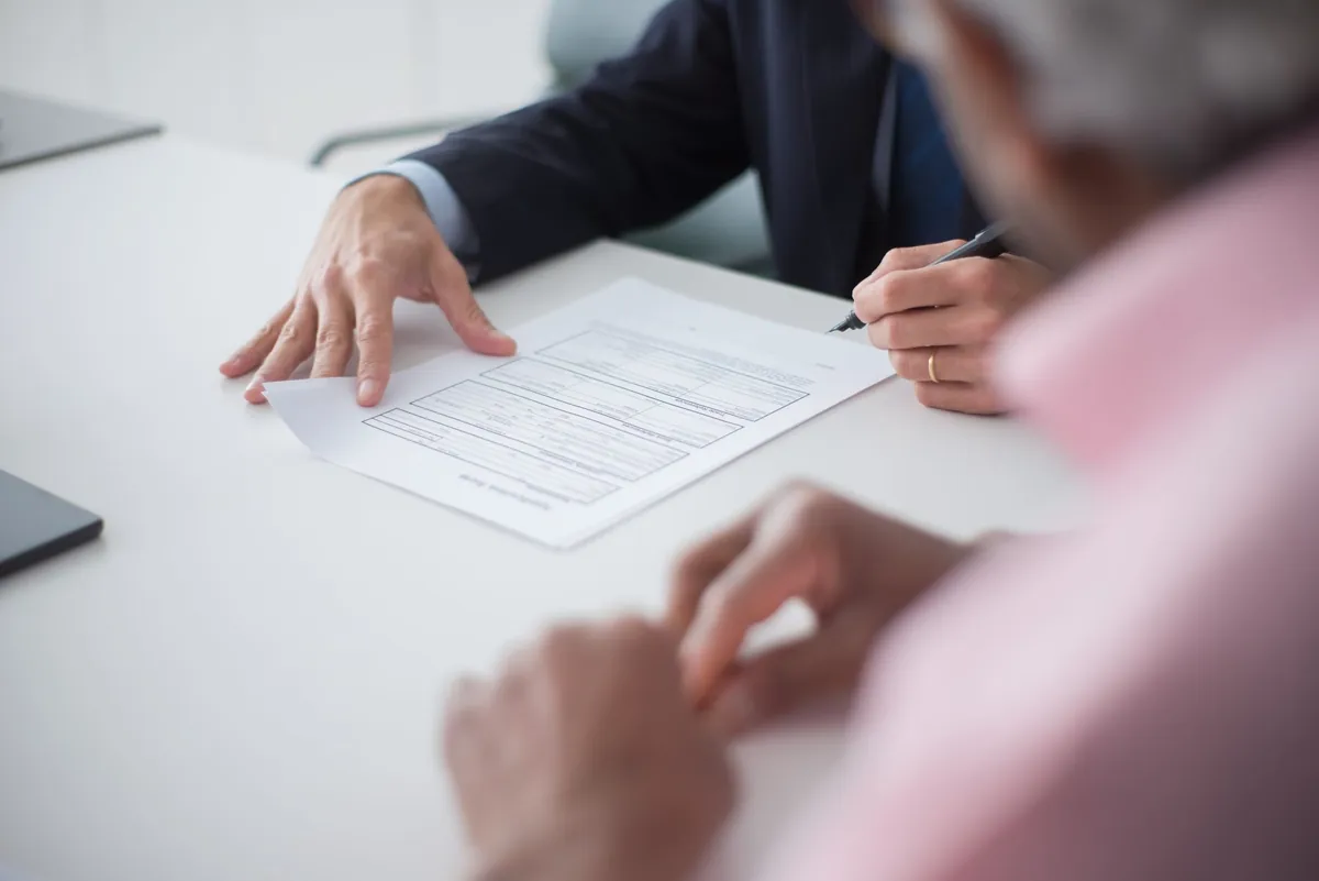 Two professionals in business attire shaking hands over a signed consulting agreement during a meeting.