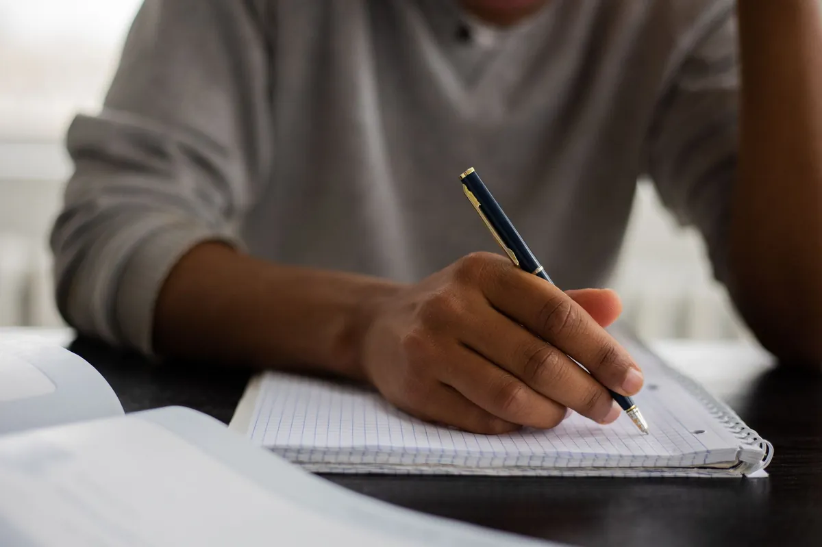 African American male student writing notes in a workbook at a table with an open textbook, blurred background.