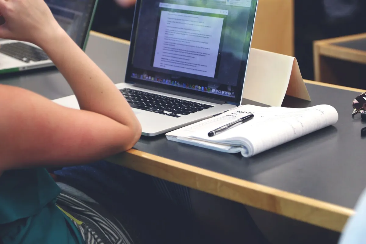A student in a classroom using a laptop while taking handwritten notes in a notebook.